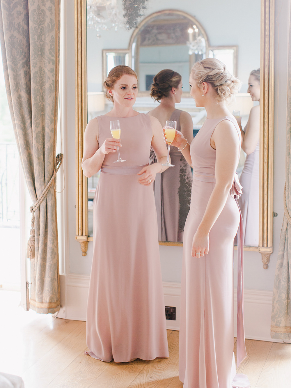 Bridesmaids stand in front of gold vintage mirror with champagne glasses at Rockbeare Manor